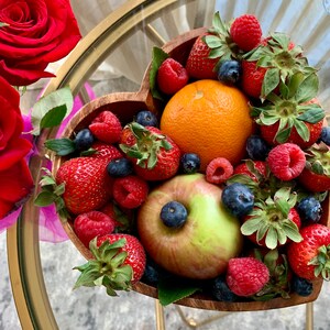 May include: A heart-shaped wooden bowl filled with fresh strawberries, raspberries, blueberries, an orange, and a green apple. The bowl sits on a gold-colored table with a glass top. Red roses are visible in the background.