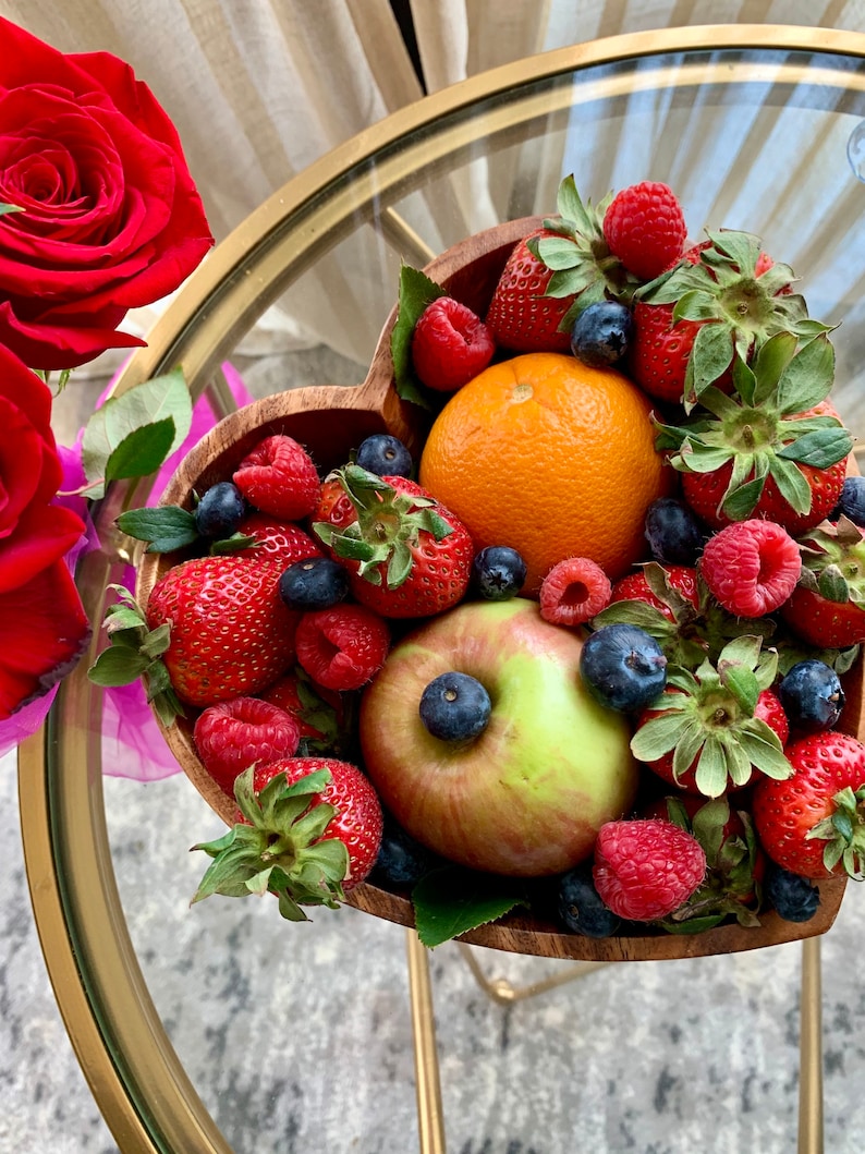 May include: A heart-shaped wooden bowl filled with fresh strawberries, raspberries, blueberries, an orange, and a green apple. The bowl sits on a gold-colored table with a glass top. Red roses are visible in the background.