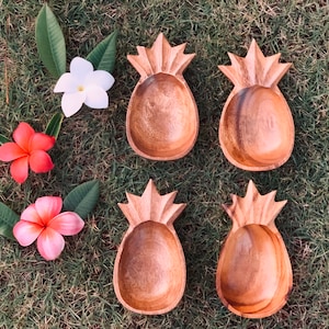Könnte beinhalten: Vier holzgeschnitzte Schalen in Ananasform mit natürlicher Holzoberfläche liegen auf grünem Gras. Die Schalen sind von grünen Blättern und bunten Blumen umgeben, darunter weiße, rosa und orangefarbene Blüten.