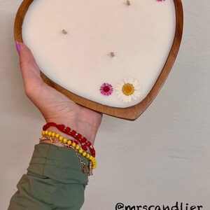 May include: Heart-shaped candle in a wooden holder. The white candle is decorated with dried flowers in pink, yellow, and white. The candle has three wicks. The text "@mrscandlier" is in the bottom right corner.