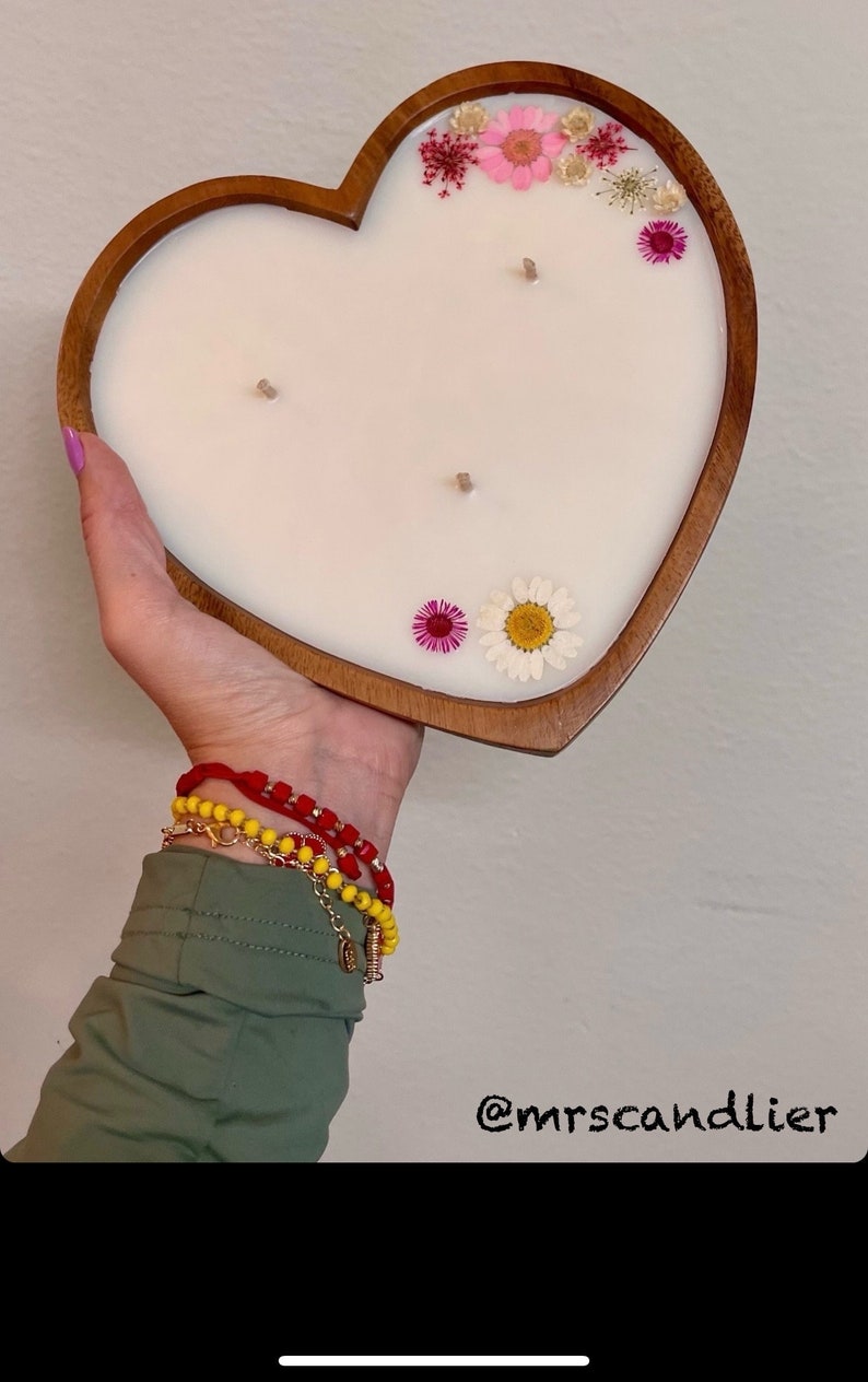 May include: Heart-shaped candle in a wooden holder. The white candle is decorated with dried flowers in pink, yellow, and white. The candle has three wicks. The text "@mrscandlier" is in the bottom right corner.