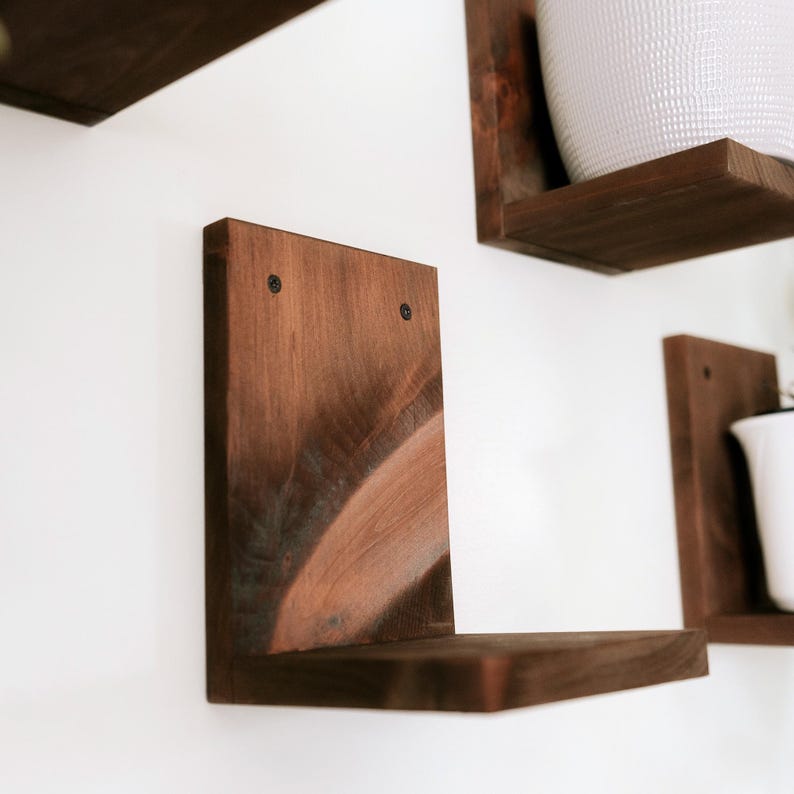 May include: Wooden floating shelves in a warm brown stain, mounted on a white wall. The shelves are L-shaped, with visible wood grain and dark screws. The design is minimalist and suitable for displaying plants or decor.