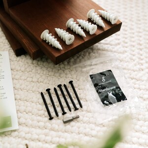 May include: A hardware kit with white plastic wall anchors, black screws, and a small tool. The kit includes a clear plastic bag with the words "Hardware Kit" printed on it. The items are arranged on a dark brown wooden surface.