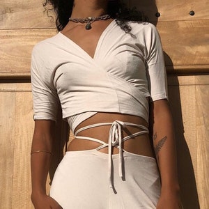 May include: A cream-coloured two-piece outfit. The top is a wrap-style crop top with short sleeves. The bottoms are high-waisted, wide-leg trousers. The outfit is made of a soft, flowing fabric. The model is standing in front of a wooden door.
