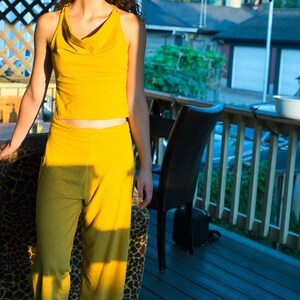 Pode incluir: Uma mulher vestindo um top amarelo sem mangas e cal&ccedil;as combinando est&aacute; em p&eacute; em um deck de madeira. Ela est&aacute; olhando para o lado e o sol est&aacute; brilhando sobre ela.