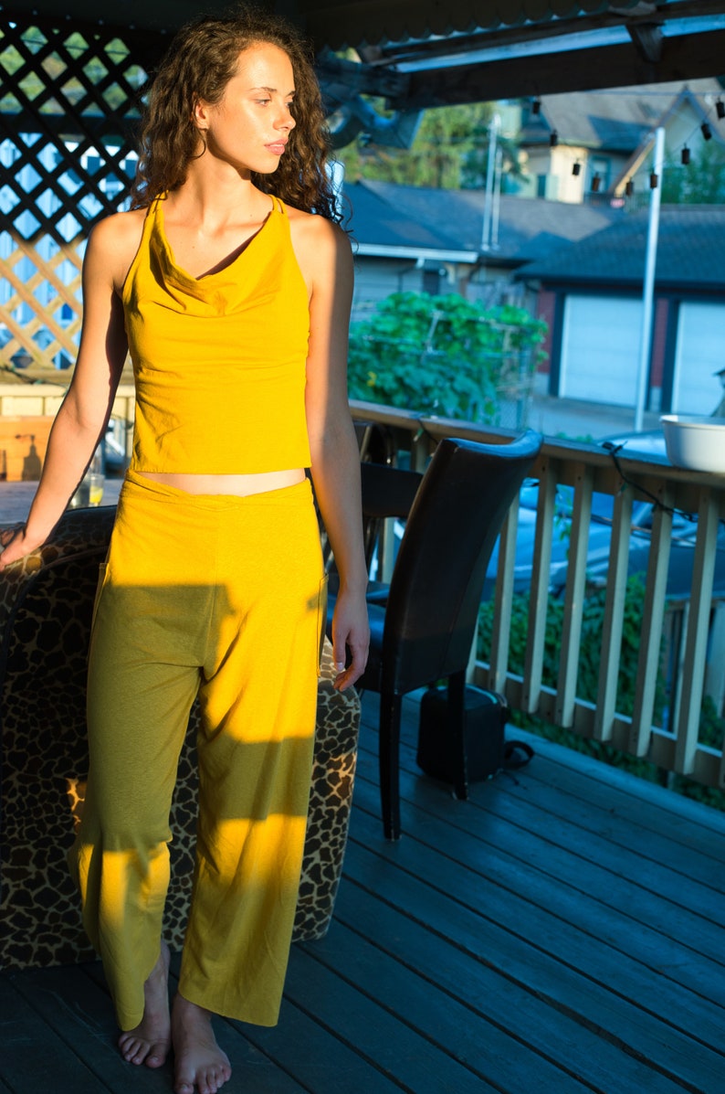 Pode incluir: Uma mulher vestindo um top amarelo sem mangas e cal&ccedil;as combinando est&aacute; em p&eacute; em um deck de madeira. Ela est&aacute; olhando para o lado e o sol est&aacute; brilhando sobre ela.