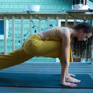 Pode incluir: Uma pessoa em um macac&atilde;o amarelo realiza uma pose de ioga em um tapete de ioga preto em um deck de madeira. A pessoa est&aacute; voltada para o lado esquerdo da imagem e tem cabelos longos e cacheados. O deck tem uma cerca de madeira com luzes de corda penduradas.