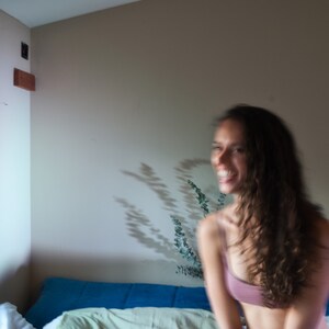 May include: A person wearing a pink top smiles in a room with a window and a blue couch. The room has a neutral wall colour and a shadow of a plant is visible on the wall.