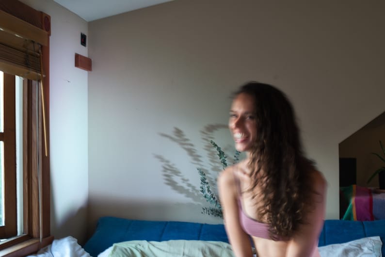 May include: A person wearing a pink top smiles in a room with a window and a blue couch. The room has a neutral wall colour and a shadow of a plant is visible on the wall.