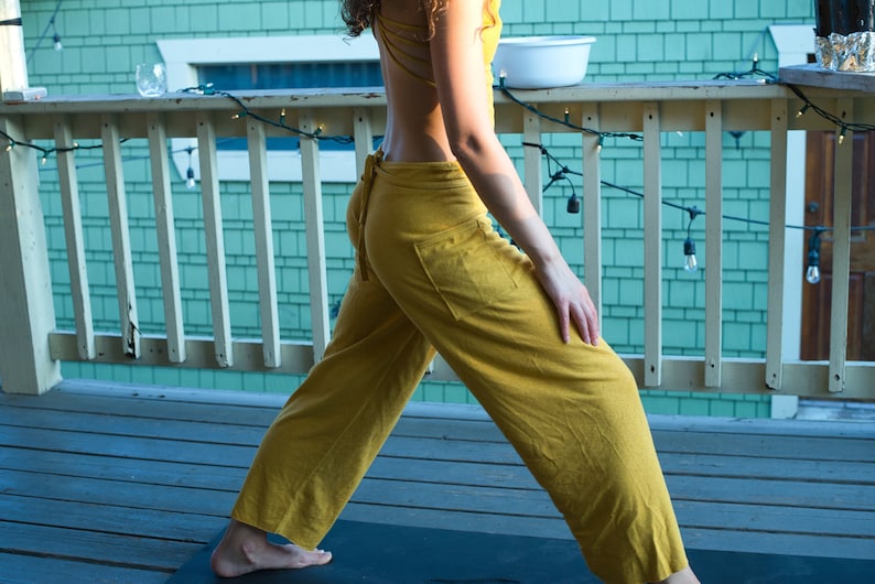 Pode incluir: Uma pessoa vestindo um macac&atilde;o amarelo mostarda com costas baixas, em p&eacute; em um deck de madeira com uma cerca e luzes de corda.