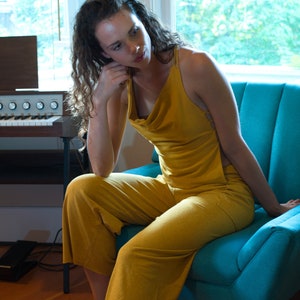 Pode incluir: Uma mulher vestindo um macac&atilde;o amarelo mostarda senta-se em uma poltrona azul turquesa. A poltrona tem um encosto estofado e est&aacute; em uma sala com um &oacute;rg&atilde;o vintage.