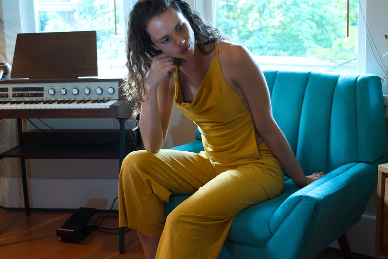 Pode incluir: Uma mulher vestindo um macac&atilde;o amarelo mostarda senta-se em uma poltrona azul turquesa. A poltrona tem um encosto estofado e est&aacute; em uma sala com um &oacute;rg&atilde;o vintage.