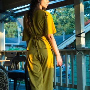 Pode incluir: Uma mulher vestindo um macac&atilde;o amarelo mostarda com um la&ccedil;o na cintura. Ela est&aacute; em p&eacute; em uma varanda de madeira com uma grade e uma vista de &aacute;rvores e casas ao fundo.