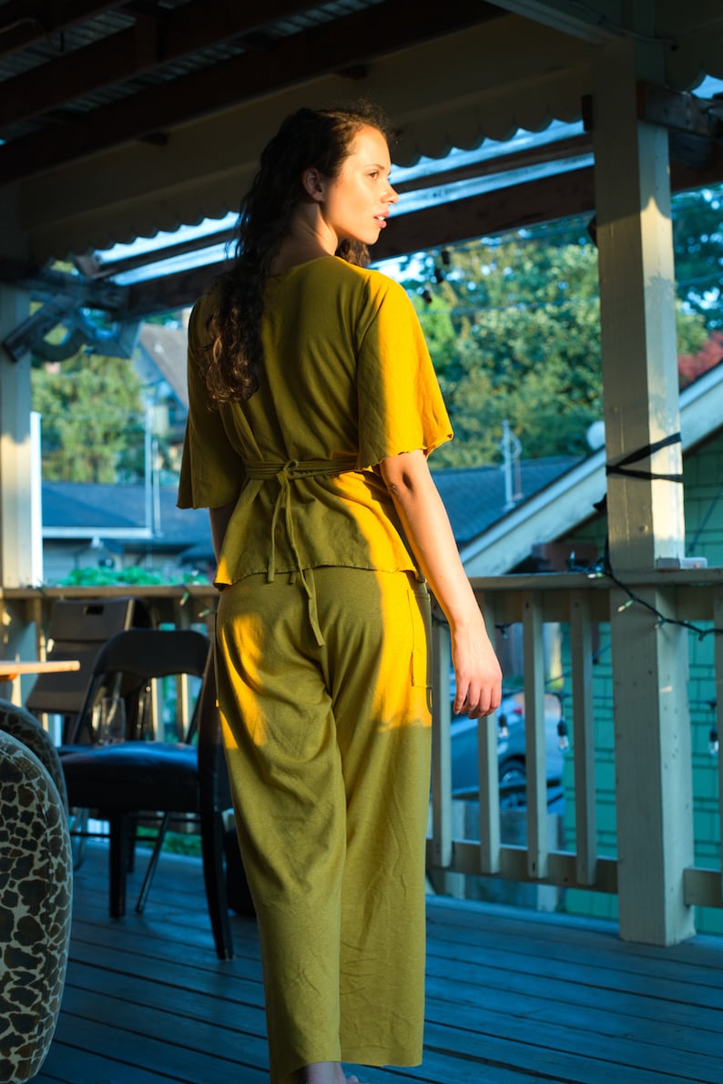 Pode incluir: Uma mulher vestindo um macac&atilde;o amarelo mostarda com um la&ccedil;o na cintura. Ela est&aacute; em p&eacute; em uma varanda de madeira com uma grade e uma vista de &aacute;rvores e casas ao fundo.