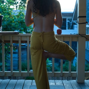 Pode incluir: Uma mulher com um macac&atilde;o amarelo est&aacute; em p&eacute; em um tapete de ioga em uma varanda de madeira. Ela est&aacute; fazendo uma pose de ioga com uma perna levantada e os bra&ccedil;os ao lado do corpo.