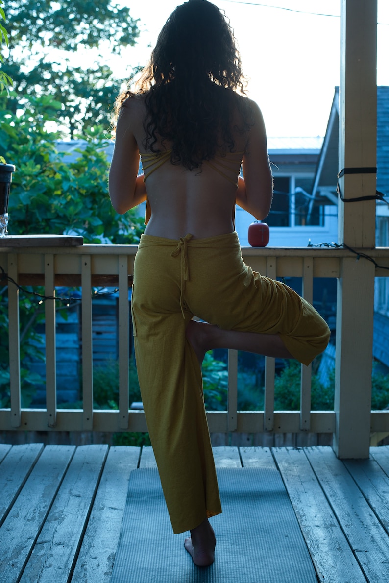 Pode incluir: Uma mulher com um macac&atilde;o amarelo est&aacute; em p&eacute; em um tapete de ioga em uma varanda de madeira. Ela est&aacute; fazendo uma pose de ioga com uma perna levantada e os bra&ccedil;os ao lado do corpo.