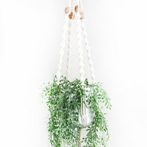Op de afbeelding: Een witte macrame plantenhanger met houten kralen hangt aan het plafond en houdt een witte pot met een groene, bladerrijke plant.