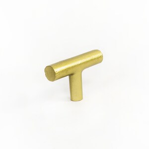 May include: A gold-coloured T-shaped cabinet knob with a textured finish.