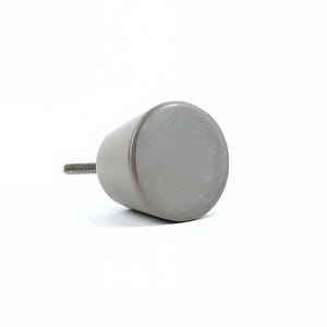 May include: A grey, cylindrical cabinet knob with a threaded screw extending from one end. The knob has a flat, circular face and a smooth, matte finish. The hardware is designed for use on drawers or cabinet doors.