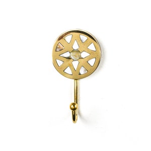 May include: A gold metal hook with a circular design featuring a geometric pattern and a white center. The hook is attached to a white wall.