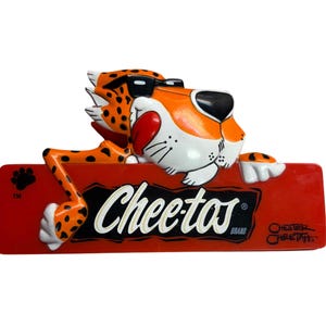 May include: A three-dimensional plastic sign featuring the orange and black spotted Chester Cheetah character wearing black sunglasses, leaning over a rectangular red plaque with the white text Cheetos Brand and the signature Chester Cheetah.