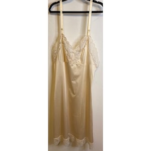 May include: A cream-colored slip dress with adjustable straps, lace trim, and a satin finish. The dress is hanging on a black hanger against a white wall. The lace trim is visible along the neckline and hem.
