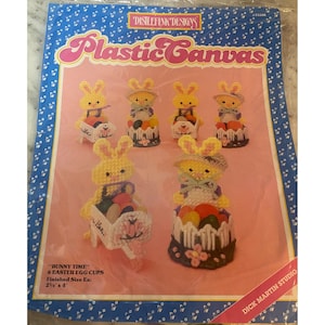 May include: A package of plastic canvas craft kit featuring six Easter egg cups with bunny designs. The bunnies are yellow and white, holding colorful eggs. The package has the text "Bunny Time" and "Plastic Canvas". Finished size is 2.25" x 4".