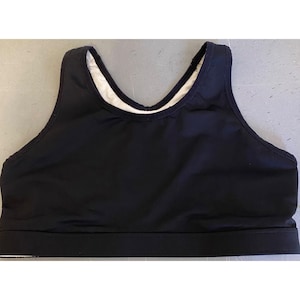 May include: A black sports bra with a scoop neckline and a white interior lining. The bra has a wide band at the bottom and a racerback design. The fabric appears to be smooth and slightly shiny.