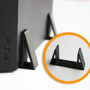 Könnte beinhalten: Schwarzer Plastikständer für eine PlayStation 4 Konsole. Der Ständer hat zwei dreieckige Stützen, die die Konsole anheben.