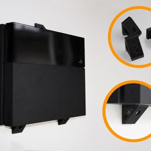 Könnte beinhalten: Schwarze Wandhalterung für eine Spielkonsole. Die Konsole wird mit schwarzen Halterungen an der Wand befestigt. Zusätzliche schwarze Halterungen sind in einem kreisförmigen Einsatz dargestellt. Die Konsole ist schwarz.