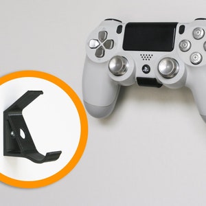 Puede incluir: Un mando de PlayStation 4 blanco y gris con detalles plateados está montado en una pared blanca. Un soporte de pared negro para el mando se muestra en un marco circular con un borde naranja.