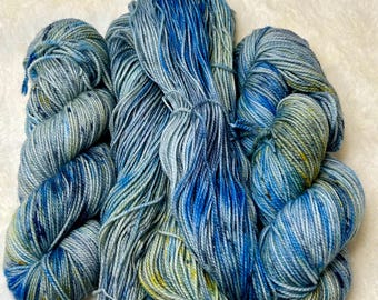 May include: A collection of hand-dyed yarn skeins in blue, teal, and yellow hues. The yarn is arranged in a loose pile, showcasing varying thicknesses and textures. The colours blend, creating a watercolour effect. The yarn is likely wool or a similar fibre, suitable for knitting or crochet.