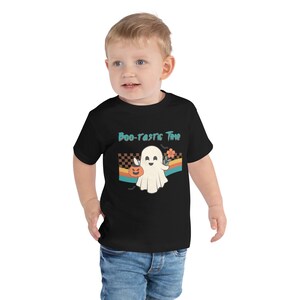 May include: A black short-sleeve t-shirt featuring a graphic of a white ghost holding an orange jack-o'-lantern and a flower. The design includes a checkered pattern, horizontal stripes, and the text "Boo-tastic Time" in teal lettering.