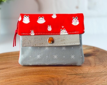 May include: A small, fabric wallet with a red flap patterned with white frog illustrations. The wallet has a gray body with a white crosshatch pattern and a light gray cork-like accent. A red cord is attached to the side.