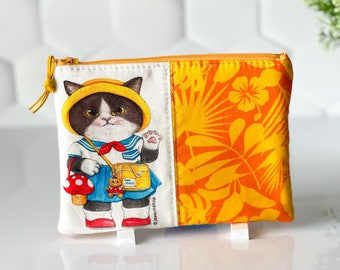 May include: A yellow and orange zippered pouch with a white panel featuring a black and white cat wearing a yellow hat and sailor outfit. The cat is holding a red and white mushroom and a small teddy bear. The pouch has a tropical print on the other side.