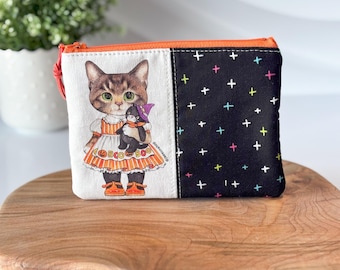 May include: A white and black zippered pouch with a cat wearing a dress and holding a penguin. The pouch has a black background with white plus signs and a white front with a cat illustration.