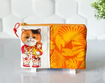 May include: A small orange and yellow zippered pouch with a white panel featuring a cartoon cat wearing overalls and holding a doll. The other side of the pouch has a tropical leaf pattern.