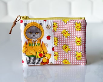 May include: A yellow and pink fabric pouch with a zipper closure. The front of the pouch features a cartoon cat wearing a yellow hoodie with the word "Kitty" on it. The back of the pouch features a pink and white checkered pattern with yellow teddy bears.