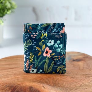 May include: A small, square fabric pouch with a dark teal background and a floral pattern. The pattern includes pink, yellow, and white flowers, along with green leaves and berries. The pouch is sitting on a wooden surface.
