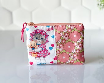 May include: A pink and white zippered pouch with a floral pattern on one side and a cartoon cat holding a doll on the other side. The pouch has the word "Rumi" printed on it.