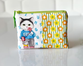 May include: A green zippered pouch with a white background. The front of the pouch features a cartoon cat wearing a blue shirt with the word "GIMU" on it. The cat is holding a lollipop and has a black and white face. The background of the pouch is a white fabric with a pattern of letters. The back of the pouch is a white fabric with a pattern of yellow and orange flowers.