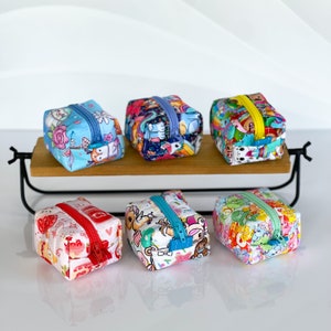 May include: Six small zippered pouches with colorful cartoon designs. The pouches are arranged in two rows of three on a wooden shelf.