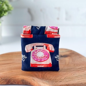 May include: A navy blue fabric pouch featuring a retro telephone design in pink and red. The pouch has a rectangular shape with a fold-over top. White floral accents are on the sides. The pouch is placed on a wooden surface.