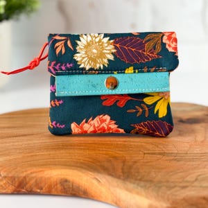 May include: A small, floral-patterned wallet with a teal accent and a snap closure. The wallet features a dark teal background with orange, yellow, and white flowers and leaves. A red cord is attached to the side.