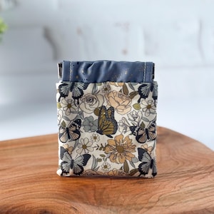 May include: A small, fabric pouch with a floral and butterfly pattern. The main body of the pouch features a cream background with butterflies, flowers, and foliage in shades of brown, black, and green. The top is a solid blue fabric.