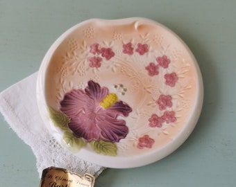 May include: A small, white ceramic dish with a pink and orange floral design. The dish features a large purple hibiscus flower with green leaves and smaller pink flowers around the edge. The dish has a gold label that reads "A. Clement France Barbotine" and "Decoré à la Main - Made in France".
