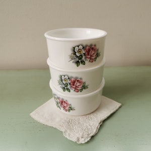 May include: Three white ceramic bowls with floral designs. Each bowl features a pink and white rose with green leaves. The bowls are stacked on top of each other.