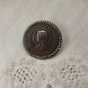 Könnte beinhalten: Silberne Brosche mit einem detaillierten Design, das das Profil einer Frau mit Schleier zeigt. Die Brosche ist rund und hat einen strukturierten Rand.