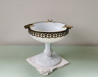 May include: White milk glass ashtray with a gold metal rim and two cigarette rests. The ashtray is on a pedestal base.