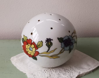 May include: White ceramic sphere with floral designs in red, yellow, and blue. The sphere has several small holes on top. The sphere is resting on a white lace cloth.